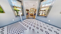Located at 165 Kathann Dr in Hampton, Virginia, Abbington's leasing office is a thoughtfully designed, single-story facility that blends modern aesthetics with practical functionality. The entrance greets visitors with a striking patterned penny-tile floor and floor-to-ceiling glass panels that flood the vestibule with natural light and offer views of the surrounding landscaped grounds.

Inside, the open lounge area features wood-look flooring, a leather sofa, and colorful accent seating arranged around a decorative open-grid room divider adorned with vibrant artwork — creating a welcoming, contemporary atmosphere. An adjacent seating nook with upholstered benches and a side door provides direct access to an outdoor patio.

The leasing area transitions into a professional workspace with patterned modular carpet tiles, light wood workstations, and modern guest chairs. Private offices feature arched transom windows and bold graphic drapery, adding character while maintaining natural light. A glass-panel door connects the main office floor to the lounge and meeting areas seamlessly.

A dedicated meeting room features bold chartreuse accent walls, a marble-top round table, and generous windows overlooking the exterior. The space also includes a café-style refreshment area with a live plant wall, pendant lighting, a high-top communal table, and a coffee station — providing a vibrant and functional hub for staff and visitors alike.