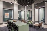 Located at 3 Hawkins Ave in Ronkonkoma, NY, these office rooms offer a thoughtfully designed, single-floor workspace that balances open collaboration with private functionality. The layout encompasses a generous footprint, providing ample room for a variety of work settings and configurations.

The open common area serves as the heart of the space, featuring a long communal bar-height table with seating, ideal for casual meetings or shared work sessions. A separate lounge zone offers comfortable chairs and a round coffee table atop a geometric-patterned area rug, encouraging informal gatherings. A dedicated refreshment station, set against a striking geometric-tiled accent wall in muted teal and white tones, is equipped with a coffee machine and counter space with cabinetry.

Glass-walled, black-framed conference rooms line the perimeter, offering visual connectivity while maintaining acoustic privacy. These enclosed rooms are outfitted with conference tables and seating for focused meetings. Secluded high-back lounge pods provide additional private nooks for individual work or small discussions.

Soaring ceilings with exposed industrial-style baffle panels and pendant lighting lend the space a contemporary, polished aesthetic. Light tile flooring transitions to patterned carpet in seating areas, adding warmth and definition. A corridor with arched architectural detailing connects to elevator access, completing this well-appointed professional environment.