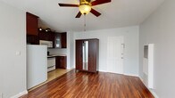 This single-floor space offers a practical and cohesive layout designed for comfortable everyday living. The main living area is an open, airy room finished with rich, warm-toned hardwood flooring that runs throughout, creating a sense of continuity and warmth. A ceiling fan with a built-in light fixture serves as a functional centerpiece overhead, while natural light filters in through windows fitted with blinds. A wall-mounted heating unit adds to the room's practicality.

The kitchen transitions seamlessly from the living area and is finished with light tile flooring. It features an abundance of deep espresso-toned cabinetry, offering generous storage above and below. Light-colored countertops provide ample workspace, complemented by a gas range with an over-the-range microwave, a refrigerator, and a stainless steel sink. Track lighting mounted beneath the upper cabinets adds functional illumination to the space.

The bathroom is clean and straightforward in design, featuring white tile surrounding a shower-tub combination, a classic pedestal sink, a wall-mounted mirror, and tile flooring. An elegant multi-bulb light fixture above the mirror provides warm, even lighting throughout the space.