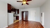 This single-floor space offers a practical and cohesive layout designed for comfortable everyday living. The main living area is an open, airy room finished with rich, warm-toned hardwood flooring that runs throughout, creating a sense of continuity and warmth. A ceiling fan with a built-in light fixture serves as a functional centerpiece overhead, while natural light filters in through windows fitted with blinds. A wall-mounted heating unit adds to the room's practicality.

The kitchen transitions seamlessly from the living area and is finished with light tile flooring. It features an abundance of deep espresso-toned cabinetry, offering generous storage above and below. Light-colored countertops provide ample workspace, complemented by a gas range with an over-the-range microwave, a refrigerator, and a stainless steel sink. Track lighting mounted beneath the upper cabinets adds functional illumination to the space.

The bathroom is clean and straightforward in design, featuring white tile surrounding a shower-tub combination, a classic pedestal sink, a wall-mounted mirror, and tile flooring. An elegant multi-bulb light fixture above the mirror provides warm, even lighting throughout the space.