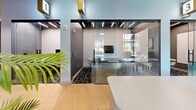 Located at 147 W Wood St in West Lafayette, Indiana, this single-floor commercial space offers a thoughtfully designed and modern work environment. The layout includes a welcoming reception area anchored by a sleek white curved desk, light hardwood flooring, and lush tropical plants that add a refreshing organic touch to the contemporary aesthetic.

Glass-walled meeting rooms line the hallways, offering transparency and an open feel while maintaining functional separation. Each meeting room is outfitted with a light wood conference table, comfortable upholstered chairs, and bold graphic accent walls featuring an artistic collage-style mural in muted gold, black, and white tones. Linear LED ceiling fixtures provide consistent, professional illumination throughout.

One of the meeting rooms features a wall-mounted flat-screen display and a built-in whiteboard, making it well-suited for presentations and collaborative sessions. The patterned dark carpet in the meeting rooms contrasts smartly with the hardwood flooring in the common areas.

The main lounge or collaboration zone benefits from large windows dressed with warm amber curtains, allowing natural light to filter in while adding a rich color accent. Open shelving with greenery and stylish pendant lighting complete the inviting atmosphere. Numbered room identifiers with gold accents add a refined, organized touch to the overall design.