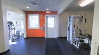 This single-floor fitness facility is designed to provide a functional and motivating workout environment. The space is characterized by its durable dark rubber flooring, which spans the exercise area and provides both safety and comfort underfoot during workouts. Vibrant orange accent walls energize the atmosphere, creating a dynamic visual contrast against the neutral white ceilings and trim throughout the room. Recessed lighting in the ceiling, complemented by natural light streaming through windows, ensures the space is well-lit at all times. The room features a white paneled exit door with glass panes, allowing additional daylight to filter in while maintaining clear egress. Strength training equipment, including a multi-function cable or weight machine, is positioned along one side of the room, offering users a range of resistance exercise options. The layout is open and spacious, allowing for free movement between equipment and exercise zones. Wall-mounted controls and fixtures suggest the room is equipped with climate or security management systems, contributing to a comfortable and secure workout setting. Overall, the facility presents a clean, purpose-built space suited for strength and general fitness training.