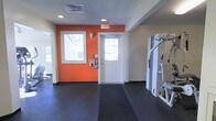 This single-floor fitness facility is designed to provide a functional and motivating workout environment. The space is characterized by its durable dark rubber flooring, which spans the exercise area and provides both safety and comfort underfoot during workouts. Vibrant orange accent walls energize the atmosphere, creating a dynamic visual contrast against the neutral white ceilings and trim throughout the room. Recessed lighting in the ceiling, complemented by natural light streaming through windows, ensures the space is well-lit at all times. The room features a white paneled exit door with glass panes, allowing additional daylight to filter in while maintaining clear egress. Strength training equipment, including a multi-function cable or weight machine, is positioned along one side of the room, offering users a range of resistance exercise options. The layout is open and spacious, allowing for free movement between equipment and exercise zones. Wall-mounted controls and fixtures suggest the room is equipped with climate or security management systems, contributing to a comfortable and secure workout setting. Overall, the facility presents a clean, purpose-built space suited for strength and general fitness training.