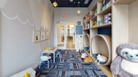 The Grove Children's Room in Piscataway, NJ, is a dedicated play space crafted with young children in mind, combining imaginative design with functional amenities. The room features soaring ceilings painted in a deep teal blue, which contrast beautifully with the light gray walls adorned with painted mountain silhouettes and framed animal artwork, creating a whimsical, storybook ambiance throughout.

A standout element is the custom-built light wood shelving unit, which incorporates circular tunnel nooks padded with tufted blue cushions — ideal cozy retreats for small children. The shelving also provides generous storage through open cubbies filled with colorful bins, books, and stuffed animals. Adjacent to this feature wall, a built-in bench with plush blue cushioning sits beneath a wall-mounted flat-screen television, offering a comfortable media viewing area.

Large windows fitted with white shutters invite ample natural light into the space, enhancing its bright and cheerful atmosphere. The patterned blue and gray modular carpet tiles ground the room with color and texture, complementing the overall design palette. A soft seating lounge area, child-sized activity tables and chairs, and open floor space provide multiple zones for play, creativity, and relaxation, making this a versatile and engaging environment for children.