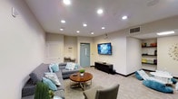 This single-floor space functions as a well-appointed communal lounge, designed to provide a comfortable and inviting environment for socializing and relaxation. The room features neutral-toned walls and light carpeted flooring, creating a clean and understated backdrop that complements the contemporary furnishings throughout.

Seating is generously provided by a large sectional sofa in grey upholstery, accented with teal and patterned throw pillows that introduce a pop of color to the space. Additional grey accent chairs contribute to a cohesive and modern aesthetic. A round wooden coffee table with a metallic base serves as the centerpiece of the seating arrangement, offering both style and functionality.

A wall-mounted flat-screen television is positioned prominently on one wall, supported by a dark media console beneath it, making the space well-suited for group viewing. Recessed ceiling lights are distributed evenly throughout, providing bright, consistent illumination across the room.

A teal-colored door with a narrow window adds a subtle architectural detail and likely leads to an adjacent corridor or exit. The overall ambiance is clean, modern, and purposefully arranged to encourage community interaction.