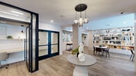 Located at 169 Johnson Rd in Morris Plains, NJ, this single-floor community space offers a generously sized, open-concept layout that seamlessly blends a living room and dining area into one cohesive environment. The living area is anchored by a striking slatted wood feature wall housing a large flat-screen TV and a stone-clad fireplace, creating a warm and inviting focal point. Curated seating — including plush swivel chairs, a soft sectional, and marble-topped coffee tables — is arranged across a patterned area rug, while an elegant pendant light cluster adds a refined ambiance overhead. An open shelving unit serves as a natural divider, styled with decorative objects that contribute to the space's contemporary aesthetic. The dining area is bright and airy, bathed in natural light from oversized windows and glass French doors that open directly to a landscaped outdoor patio. Light hardwood flooring runs throughout, complementing the warm, neutral color palette. Additional seating nooks with bistro-style high-top tables offer flexible use. A bar area with stone tile accents and bar seating adds further functionality. The overall design balances modern sophistication with approachable comfort.
