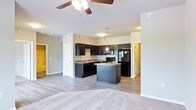 This single-floor residence offers a thoughtfully designed open-concept layout that connects the kitchen and living areas in a seamless, functional flow. The kitchen is a standout feature, showcasing dark espresso cabinetry that extends to the upper walls, complemented by granite-style countertops that provide both durability and visual appeal. A central kitchen island adds valuable workspace and serves as a natural gathering point. Black appliances, including a refrigerator and range, lend a cohesive, modern aesthetic to the space. Pendant lighting above the island and recessed lighting throughout ensure the kitchen is well-lit and inviting. The adjoining living area transitions smoothly with warm hardwood-style flooring that gives way to plush carpeting, creating distinct zones within the open plan. A ceiling fan with a light fixture enhances comfort and airflow in the living space. Neutral gray wall tones provide a versatile backdrop that complements the darker cabinetry and flooring. Doorways visible from the main area suggest additional rooms, including what appears to be a closet or utility space and a hallway leading to further living quarters, making this a well-connected and practical single-level home.