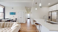 This thoughtfully designed single-floor residence showcases a clean, contemporary aesthetic defined by crisp white walls, warm hardwood flooring, and an abundance of natural light. The open-concept living and kitchen area serves as the heart of the home, where a sleek peninsula with a white quartz countertop and brass-accented bar stools connects the kitchen to the living space. The kitchen is equipped with stainless steel appliances, white shaker cabinetry, and pendant lighting that adds a modern touch. The living area features large, black-framed windows that frame urban cityscape views and flood the room with daylight, complemented by a coffered ceiling detail that adds architectural interest. A hallway leads to two well-proportioned bedrooms, each offering hardwood floors, generous windows with city views, and closet storage. The primary bedroom includes a spacious closet with rod and shelf organizers. The bathroom is elegantly finished with marble-look tile flooring, white subway tile surrounding a soaking tub with glass enclosure, and a rainfall showerhead. Throughout the residence, recessed lighting, clean lines, and a neutral palette create a cohesive, modern living environment.