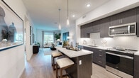 Located at 6000 Luminary Drive in Chula Vista, CA, this thoughtfully designed one-bedroom, three-bathroom residence offers a modern living experience across a single floor. The open-concept layout seamlessly connects the kitchen, dining, and living areas, creating an inviting and functional space bathed in natural light from large windows with views of swaying palm trees.

The contemporary kitchen is a standout feature, boasting rich dark cabinetry, white quartz countertops, a subway tile backsplash, stainless steel appliances, and a central island with pendant lighting and bar seating — perfect for casual dining or entertaining. Light wood-style flooring flows throughout, complementing the clean, neutral palette.

The spacious primary bedroom features a ceiling fan, large window, accent wallcovering, and ample closet storage complete with built-in organization rods. The three bathrooms maintain the home's sleek aesthetic with dark wood vanities, white countertops, and tub-shower combinations.

Additional highlights include a dedicated laundry area, multiple closets for generous storage, a private balcony, and well-connected hallways. The bright living room offers a comfortable retreat with garden views, while the overall design strikes a balance between modern sophistication and everyday livability.
