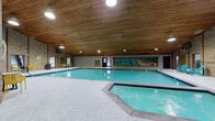 This single-story indoor aquatic facility is designed to provide a welcoming and functional environment for swimmers and leisure guests alike. The centerpiece of the space is a large, sparkling swimming pool with clear turquoise water, accompanied by a separate hot tub area positioned at one end. The pool deck offers ample room to move around, furnished with colorful Adirondack chairs in teal and casual seating sets in yellow, creating relaxed gathering spots throughout.

Architecturally, the facility is defined by its striking warm-toned wood-planked ceiling, which lends a rustic, cabin-like character to the interior. The exposed brick walls reinforce this aesthetic, while large windows along the perimeter invite natural light and provide views of the surrounding greenery outdoors. A decorative mural depicting a sunny outdoor pool scene adds a vibrant, artistic focal point along one wall.

Access points include a distinctive diagonal wood-paneled door and glass double doors framed by additional brick detail. A utility area near the entrance houses a vending machine and swim class equipment, indicating the facility's use for organized aquatic programs. Recessed and pendant lighting supplements the natural light throughout. The overall atmosphere is functional yet inviting, suited for recreational swimming, aquatic instruction, and group leisure activities.