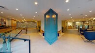 This elegantly designed lobby makes an immediate impression with its striking teal architectural column at the center of the space, adorned with brass wall sconces that cast a warm, inviting glow. The open-plan layout is organized across split levels connected by wide, shallow staircases with sleek metal and glass railings, creating a dynamic flow throughout the space.

Multiple distinct seating areas are thoughtfully distributed across the lobby. One lounge features a linear electric fireplace set against a dramatic marble-effect wall, paired with mustard velvet armchairs, navy blue ottomans, and a colorful framed artwork above. Another lounge area offers modern egg-style chairs in deep navy arranged around a solid wood coffee table, with a large-scale art piece as a focal point against a textured dark wall panel.

Natural light filters in through floor-to-ceiling windows overlooking a lush green courtyard, adding a refreshing outdoor connection to the interior. Warm wood paneling and cabinetry appear throughout, complemented by large-format light stone flooring. Curated sculptures and decorative objects are displayed on a built-in credenza with a fluted white accent wall backdrop.

The elevator bank features wood-wrapped surrounds with stainless steel doors, accented by the recurring teal and brass detailing. A dedicated workspace nook and what appears to be a concierge or service counter complete the functional offerings of this polished, art-forward lobby.