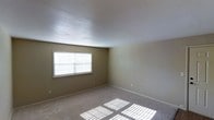 This single-floor space presents a clean and functional layout designed with simplicity in mind. The interior features neutral-toned walls that create a calm, understated atmosphere throughout. The room is finished with wall-to-wall carpet in a light, neutral shade, providing a soft and comfortable underfoot feel that complements the overall aesthetic. A textured ceiling adds subtle visual interest overhead, while white baseboards frame the perimeter of the room with a crisp, finished look. A prominently positioned window fitted with horizontal blinds allows generous natural light to flow into the space, casting defined light patterns across the carpeted floor and contributing to an open, airy ambiance. The walls are smooth and move-in ready, offering a versatile blank canvas for a variety of uses and furnishing styles. Electrical outlets are conveniently placed along the walls, ensuring practical functionality. The overall design is straightforward and understated, making it an adaptable space well-suited for comfortable everyday living.