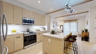 This thoughtfully designed single-floor residence at 82–98 Jolley Dr in Bloomfield, Connecticut, blends modern aesthetics with everyday functionality. The open-concept kitchen is a standout feature, showcasing light wood cabinetry that extends to the ceiling, stainless steel appliances including a range, microwave, and refrigerator, and a generous island with a white quartz countertop and matte black fixtures. A sculptural multi-globe pendant light adds an artistic touch, while light hardwood flooring and exposed wood ceiling beams contribute warmth throughout the living and kitchen areas. The living room is bright and inviting, with sheer curtained sliding doors that bathe the space in natural light and offer outdoor access. The bedrooms are comfortable and serene, finished with plush carpet flooring, large windows dressed in patterned sheer curtains, and clean white walls that create a calm, restful atmosphere. The bathroom features floor-to-ceiling white subway tile in the tub-shower combo, marble-look tile flooring, and matte black hardware and fixtures for a crisp, contemporary contrast. A dedicated laundry area with a front-load Samsung washer adds practical convenience to this well-appointed home.