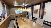 Located in Little Elm, this single-floor professional meeting space is thoughtfully designed to support focused collaboration and executive-level gatherings. The centerpiece of the room is a broad, warm-toned wood conference table surrounded by sleek, padded gray chairs that offer both comfort and a clean aesthetic. Overhead, a striking multi-shade chandelier serves as a bold design statement, casting a warm, inviting glow throughout the space. A wall-mounted flat-screen display is positioned prominently at one end of the table, providing seamless support for presentations and media sharing. The room features large black-framed windows that allow natural light to filter in while maintaining a sense of privacy, and contemporary abstract artwork adorns the walls, adding a layer of visual sophistication. Recessed lighting in the tray ceiling complements the ambient warmth of the chandelier, ensuring the space is well-lit for any occasion. A decorative planter arrangement at the center of the table introduces a touch of organic freshness to the otherwise structured, professional environment. The overall ambiance is refined yet approachable, making this space well-suited for business meetings, presentations, or team discussions.