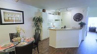 This single-floor residence showcases an open-concept design that seamlessly connects the kitchen and dining areas into one cohesive living space. The kitchen is outfitted with crisp white cabinetry extending to the ceiling, offering generous storage capacity, and is equipped with essential appliances including a refrigerator, microwave, and cooktop. A peninsula-style breakfast bar serves as a natural divider between the kitchen and the surrounding living space, providing both additional prep space and casual seating opportunities. Track lighting installed along the kitchen ceiling ensures ample illumination over the work areas. Adjacent to the kitchen, the dining area comfortably accommodates a round table with seating for multiple guests, anchored by warm hardwood-style flooring that flows consistently throughout the space. Crown molding along the upper walls adds a refined architectural touch, contributing to the clean, polished aesthetic of the interiors. The predominantly white walls and cabinetry create a bright, airy atmosphere, further enhanced by decorative touches such as a tall indoor plant and café-style signage that lend the space a welcoming, casual charm.