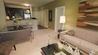 This single-floor residence offers a comfortable and cohesive living experience with a carefully considered layout and warm, inviting aesthetic. The open-concept design seamlessly connects the living area to the kitchen, creating a spacious and functional flow throughout the home. The living room features plush carpeting, olive-green accent walls, and ample room for stylish furnishings, including a glass coffee table and a cozy sofa. A sleek floor lamp adds warmth to the space, while the neutral color palette enhances the sense of openness. The kitchen is accessible via a convenient pass-through counter, offering a modern setup with dark cabinetry and essential appliances, making it ideal for both everyday cooking and entertaining. A compact dining area with a glass-top table and contemporary chairs sits at the intersection of the kitchen and living space, perfect for casual meals. A hallway leads to additional rooms, providing a clear separation between the living and private areas of the home. White trim and doors throughout contribute to a clean, polished look, while the overall design balances modern style with everyday comfort.