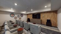 Located at 6150 Canoga Ave in the Woodland Hills neighborhood of Los Angeles, this single-floor screening room is thoughtfully designed to deliver a refined communal viewing experience. The focal point of the space is a large flat-screen television mounted on a striking textured wood accent wall, flanked by elegant wall sconces that provide warm ambient lighting. A sleek, low-profile media cabinet sits beneath the display, offering a clean and organized presentation. Rows of generously sized, upholstered lounge chairs in a neutral gray fabric are arranged in a theater-style layout, each paired with a compact round side table in a rich mahogany finish for added convenience. The opposite side of the room features floor-to-ceiling sheer drape panels covering wide windows, allowing diffused natural light to softly illuminate the space while maintaining a cinematic atmosphere. Colorful circular wall art pieces add a vibrant, contemporary touch to the neutral-toned walls, lending personality and visual interest to the room. Recessed ceiling lighting and a centralized HVAC system ensure comfort throughout. The mixed flooring—combining light and dark tones—grounds the room with a modern aesthetic. Overall, this screening room strikes a balance between functionality and sophisticated design.