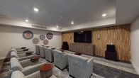 Located at 6150 Canoga Ave in the Woodland Hills neighborhood of Los Angeles, this single-floor screening room is thoughtfully designed to deliver a refined communal viewing experience. The focal point of the space is a large flat-screen television mounted on a striking textured wood accent wall, flanked by elegant wall sconces that provide warm ambient lighting. A sleek, low-profile media cabinet sits beneath the display, offering a clean and organized presentation. Rows of generously sized, upholstered lounge chairs in a neutral gray fabric are arranged in a theater-style layout, each paired with a compact round side table in a rich mahogany finish for added convenience. The opposite side of the room features floor-to-ceiling sheer drape panels covering wide windows, allowing diffused natural light to softly illuminate the space while maintaining a cinematic atmosphere. Colorful circular wall art pieces add a vibrant, contemporary touch to the neutral-toned walls, lending personality and visual interest to the room. Recessed ceiling lighting and a centralized HVAC system ensure comfort throughout. The mixed flooring—combining light and dark tones—grounds the room with a modern aesthetic. Overall, this screening room strikes a balance between functionality and sophisticated design.
