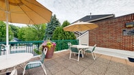 Located at 1150 Northwood Dr in Eagan, Minnesota, this outdoor sun deck offers a well-appointed rooftop retreat designed for relaxation and social gatherings. The deck features a textured, tile-paved surface that provides both durability and visual appeal across its generously sized open-air footprint.

The space is divided into two functional zones. A dedicated lounging area is furnished with multiple reclining chaise lounge chairs, allowing residents to enjoy fresh air and open skies in comfort. The dining and gathering area features round tables paired with chairs and shaded by large canopy umbrellas, making it suitable for outdoor meals or casual socializing. Vibrant potted floral arrangements add color and a welcoming touch throughout the deck.

Sturdy green metal railings enclose the perimeter of the entire deck, ensuring safety while maintaining open sightlines to the surrounding landscape. Views from the deck encompass mature evergreen and deciduous trees, as well as the neighboring building structures, creating a pleasant natural backdrop.

The deck is flanked by the building's brick and siding exterior walls, with signage and informational notices posted near the access entry. The open overhead exposure provides ample natural light throughout the day. Overall, the sun deck serves as a well-maintained communal outdoor amenity, thoughtfully designed for both leisure and social use.