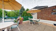 Located at 1150 Northwood Dr in Eagan, Minnesota, this outdoor sun deck offers a well-appointed rooftop retreat designed for relaxation and social gatherings. The deck features a textured, tile-paved surface that provides both durability and visual appeal across its generously sized open-air footprint.

The space is divided into two functional zones. A dedicated lounging area is furnished with multiple reclining chaise lounge chairs, allowing residents to enjoy fresh air and open skies in comfort. The dining and gathering area features round tables paired with chairs and shaded by large canopy umbrellas, making it suitable for outdoor meals or casual socializing. Vibrant potted floral arrangements add color and a welcoming touch throughout the deck.

Sturdy green metal railings enclose the perimeter of the entire deck, ensuring safety while maintaining open sightlines to the surrounding landscape. Views from the deck encompass mature evergreen and deciduous trees, as well as the neighboring building structures, creating a pleasant natural backdrop.

The deck is flanked by the building's brick and siding exterior walls, with signage and informational notices posted near the access entry. The open overhead exposure provides ample natural light throughout the day. Overall, the sun deck serves as a well-maintained communal outdoor amenity, thoughtfully designed for both leisure and social use.