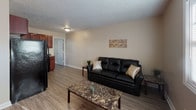 Located at 121 Harbor Pointe Ln in Greenville, North Carolina, this single-floor unit offers a practical and comfortable layout with consistent wood-look flooring running throughout the space. The living area is open and welcoming, featuring neutral beige walls, a textured ceiling, and natural light from a window fitted with blinds. The kitchen is well-appointed with rich cherry wood cabinetry, dark granite-style countertops, a stainless steel sink with under-cabinet lighting, and black appliances including a refrigerator and gas range. The layout from the aerial view reveals a connected open-plan living and kitchen area that flows efficiently into the rest of the unit. The bedrooms are comfortably sized with the same warm wood-look flooring, textured ceilings, and neutral wall tones. One bedroom features a sliding door closet and access to natural light through vertical blinds, while another bedroom offers a standard door entry and ample wall space. The bathroom includes a full-size soaking tub and shower combination with clean white surround walls and a chrome towel bar. Overall, the unit presents a clean, functional design with cohesive finishes throughout, suited for everyday living.