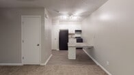 This single-floor unit at 3551 Forestdale Dr in Burlington, North Carolina, offers a practical and well-appointed layout designed for comfortable everyday living. The main living area greets residents upon entry through a classic six-panel front door, featuring plush carpeting underfoot, neutral-toned walls, and a ceiling fan with integrated lighting. A wall-mounted PTAC unit provides climate control, while a window dressed with blinds allows natural light to filter in. The space transitions seamlessly into the kitchen and dining area, where wood-look hard flooring takes over for easy maintenance. The kitchen is equipped with white cabinetry, a gas range, a full-size refrigerator, and a single-basin sink, all finished with a textured ceiling overhead and illuminated by a track lighting fixture. A peninsula with a granite-style countertop extends into the living space, offering additional prep area and casual seating. A full bathroom completes the unit, featuring a combined tub and shower with chrome fixtures and a clean, simple aesthetic. Throughout the space, consistent neutral tones, white trim, and functional finishes create a cohesive and move-in-ready environment.