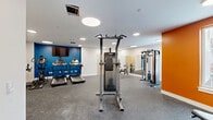Located at 431 Church St in Norfolk, Virginia, this fitness center offers a well-equipped, single-floor exercise space designed to accommodate a comprehensive range of workout routines. The room features a durable speckled rubber flooring throughout, providing both safety and comfort during exercise. Recessed circular ceiling lights illuminate the space evenly, creating a bright and energizing atmosphere.

The layout is thoughtfully organized, with cardio equipment—including multiple treadmills and stationary bikes—positioned along one side of the room. Strength training is well-represented, with a cable machine system, a pull-up and dip station, and additional functional training equipment. A large wall-mounted mirror expands the sense of space and allows users to monitor their form during workouts. Stability balls and exercise mats round out the available equipment.

Visually, the room benefits from bold accent walls in deep blue and vibrant orange, adding energy and character to the otherwise clean white interior. Natural light filters in through windows on the orange accent wall side, offering views of the surrounding area. Wall-mounted air conditioning units ensure climate comfort year-round. A wall-mounted flat-screen television is positioned near the cardio area for entertainment during workouts. The space is accessible via light wood-toned doors and maintains a clean, modern aesthetic throughout.