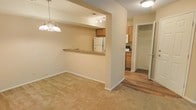 This single-floor residence offers an open and functional layout with a warm, neutral color palette throughout. The living area is finished with plush carpeting and is anchored by an elegant chandelier that adds a touch of classic style to the space. A pass-through counter with a granite-style surface connects the living area to the kitchen, creating an open sightline that enhances the sense of space.

The kitchen is equipped with light wood cabinetry, a white refrigerator, and additional appliances, all illuminated by track lighting on the ceiling. The kitchen transitions into a hallway finished with hardwood-style flooring, which leads further into the unit. A doorway visible at the end of the hallway opens to what appears to be a walk-in closet or additional storage, suggesting thoughtful use of space.

The overall ambiance is bright and functional, with recessed lighting and flush-mount ceiling fixtures providing consistent illumination. Neutral beige walls and warm-toned finishes create a cohesive and inviting atmosphere throughout the space.