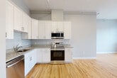This well-appointed one-bedroom, one-bathroom unit in Danvers, MA offers a thoughtfully designed open-concept layout that maximizes the generous living space. The kitchen is a standout feature, showcasing crisp white shaker cabinetry, granite countertops, a full suite of stainless steel appliances including a French door refrigerator, electric range, built-in microwave, and dishwasher. The kitchen flows seamlessly into the expansive living area, both finished with warm, light-toned hardwood flooring that enhances the bright, airy atmosphere created by recessed lighting and exposed structural columns that add an industrial character to the space. The bedroom is carpeted for comfort and features large multi-pane windows that flood the room with natural light and offer views of the surrounding neighborhood. Additional closets throughout provide ample storage. The bathroom is finished with tile flooring, a granite-topped vanity, and a shower-tub combination. A dedicated laundry area and multiple hallways contribute to the unit's functional layout. Neutral gray walls throughout create a clean, modern aesthetic that complements the unit's contemporary finishes.