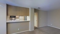 This single-floor space features an open and functional layout designed to maximize usability. The kitchen is separated from the living area by a half-wall pass-through counter, creating a connected yet defined cooking space. Upper cabinetry finished in warm wood tones provides ample storage, while appliances including a microwave and refrigerator are integrated into the kitchen area. The countertop pass-through allows for easy interaction between the kitchen and the adjacent living space.

The living area is carpeted in a neutral beige tone that complements the soft gray walls, creating a cohesive and understated aesthetic throughout. A ceiling-mounted HVAC vent ensures climate control throughout the space. The layout allows for versatile furniture arrangement within the main living zone.

A doorway visible from the living area leads to additional rooms, suggesting a logical and straightforward floor plan. White baseboards provide clean contrast along the walls. The overall ambiance is calm and neutral, offering a blank canvas well-suited for personalization. The space presents a practical and efficient design appropriate for everyday living.