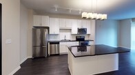 Located on Lindell Boulevard in the vibrant midtown neighborhood of St. Louis, Missouri, this one-bedroom unit offers a thoughtfully designed single-floor layout with contemporary finishes throughout. The open kitchen serves as the centerpiece of the living space, showcasing crisp white shaker cabinetry extending to the ceiling, dark granite countertops, and a full suite of stainless steel appliances including a refrigerator, electric range, microwave, and dishwasher. A classic white subway tile backsplash adds a clean, modern touch, while a kitchen island with matching granite provides additional prep and dining space. Track lighting and a modern pendant fixture illuminate the kitchen and living area, which flows toward a large window that draws in natural light and offers views of the surrounding cityscape. Rich, dark hardwood-style flooring runs throughout the main living areas, creating a warm contrast against the neutral wall tones. The bedroom is a comfortable, carpeted retreat featuring a generous window with urban views and access to a closet. The bathroom is finished with tile flooring, a white vanity with dark granite countertop, an undermount sink, and a large mirror with overhead lighting, maintaining the cohesive, polished aesthetic found throughout the unit.