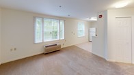 This single-floor residential unit offers a clean, functional layout designed for comfortable everyday living. The open living area is finished with neutral wall-to-wall carpeting and crisp white walls, creating a bright and airy atmosphere. Multiple windows fitted with horizontal blinds allow generous natural light to fill the space, with views of surrounding greenery. A wall-mounted HVAC unit provides individual climate control. The entry hallway leads seamlessly into the main living space, which offers ample room for a variety of furniture arrangements. The kitchen is efficiently designed in an L-shaped configuration, featuring warm oak cabinetry with substantial upper and lower storage, white laminate countertops, a stainless steel sink, and a white coil-burner electric range with overhead ventilation. The full bathroom includes tile flooring in a warm beige tone, an oak vanity with a white countertop and large mirror, and a full tub-shower combination with decorative tile accenting and accessibility grab bars. The bedroom is generously proportioned, finished with soft carpeting, a window with natural views, and its own wall HVAC unit. The overall palette of warm neutrals and natural wood tones gives the unit a cohesive, welcoming character throughout.