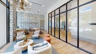 Located at 1698 Northwest 3rd Avenue in Miami's Overtown district, this thoughtfully designed lounge space offers a compelling blend of contemporary aesthetics and inviting comfort. The single-floor living area is anchored by soaring ceilings that amplify the sense of openness, while dramatic floor-to-ceiling windows framed in sleek black steel flood the interior with abundant natural light and offer clear views of the lush, tree-lined streetscape outside.

A striking geometric wallcovering — rendered in a bold, angular line pattern — serves as the room's defining design element, wrapping key walls and adding visual depth and texture throughout. Light-toned hardwood flooring runs the length of the space, complementing the bright, airy palette of white walls and natural wood furnishings.

The lounge is thoughtfully arranged with multiple distinct seating zones. Plush boucle armchairs, rattan accent chairs, and organic-shaped coffee tables are grouped atop a soft blue area rug, creating an intimate conversation area. A warm brass chandelier with candlestick-style lights serves as a refined focal point overhead. A black-framed glass partition wall adds architectural interest while defining the space without sacrificing openness. An arched wooden bookcase and curated wall art complete the sophisticated, welcoming atmosphere.