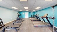 Located at 949 Lake Village Dr in Chesapeake, Virginia, this fitness center offers a dedicated exercise room designed to support a well-rounded workout routine. The space is defined by its vibrant teal-painted walls, durable rubber-composite flooring, and a drop ceiling fitted with recessed panel lighting that ensures the room is consistently and evenly illuminated throughout.

The exercise room is generously outfitted with a variety of equipment to accommodate multiple fitness goals. Cardio options include several treadmills and a stationary recumbent bike, an upright bike, and an elliptical trainer. For strength training, the room features a multi-function cable machine, a dumbbell rack stocked with a range of free weights, and adjustable weight benches. A wall-mounted split air conditioning unit provides climate control, keeping the environment comfortable year-round.

Natural light filters into the space through a set of glass double doors and flanking windows along the entry wall, offering views of the surrounding outdoor area and creating an open, airy feel. The layout is thoughtfully organized to provide clear circulation paths between equipment, allowing multiple users to exercise simultaneously without congestion. A safety defibrillator is also visibly mounted near the entrance, reflecting a commitment to user well-being.