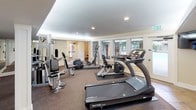 Located at 13011 Meridian E in Puyallup, WA, this dedicated fitness facility offers a comprehensive workout environment designed to accommodate a variety of exercise needs. The gym is laid out across a single floor, with distinct zones for both cardiovascular and strength training activities.

The main workout area features a combination of carpet and tile flooring, providing a practical and durable surface throughout. A row of large, multi-pane windows along one wall floods the space with natural light and offers views of the surrounding outdoor environment, creating an open and motivating atmosphere. Recessed ceiling lights and perimeter cove lighting further enhance visibility throughout the facility.

Cardio equipment includes SportsArt treadmills and elliptical machines, accompanied by a wall-mounted flat-screen television for entertainment during workouts. Strength training is well-represented with a Vectra multi-station cable machine, free weight equipment, and additional resistance machines. Floor-to-ceiling mirrors along one wall amplify the sense of space and allow users to monitor their form.

The facility also includes clearly marked restrooms for both men and women, a dedicated entry vestibule with a tiled floor and exterior glass door, and a fire safety station with an extinguisher mounted on the wall. A bulletin board near the entrance provides informational postings for users. The overall design is clean, functional, and well-maintained, making it a practical and inviting fitness resource.
