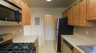 Spanning two floors, this space offers a practical and functional layout. The kitchen is designed as a galley-style corridor, maximizing efficiency with cabinetry lining both sides. Warm honey-toned wood cabinets with silver hardware provide ample storage, complemented by light-toned countertops. A black gas range sits along one wall, while a stainless-and-black refrigerator and black dishwasher complete the appliance suite. Light tile flooring runs throughout the kitchen, contributing to a clean, easy-to-maintain aesthetic. A louvered bifold door leads to an adjacent space, likely a pantry or utility area, while an electrical panel is mounted on the wall, discreetly integrated into the kitchen layout. Overhead lighting keeps the space well-lit. The kitchen connects to additional areas of the home, hinting at a broader floor plan across its two levels. Overall, the space presents a straightforward, move-in-ready environment suited for everyday living.