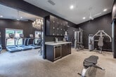 Located at 1205 Maple St in Carrollton, Georgia, this single-floor fitness center combines high-end design with comprehensive workout functionality. The space is defined by its bold aesthetic — deep matte black walls contrast sharply against light carpeted flooring and a white ceiling, creating a sleek, modern atmosphere throughout.

The main workout area is generously sized and houses a full lineup of commercial-grade cardio equipment, including multiple True-brand treadmills, elliptical trainers, a stair climber, and a cable machine. A dedicated free weights zone features a full rack of dumbbells arranged in front of a large wall mirror, which amplifies the sense of space while allowing users to monitor their form. Medicine balls and a variety of accessories are neatly organized nearby.

A frosted glass partition wall divides a secondary area, offering a semi-private space ideal for stretching, functional training, or focused workouts. Ceiling fans are installed throughout to support ventilation and comfort during exercise.

Lighting is a standout feature — recessed can lights provide even, practical illumination, while elegant crystal and white multi-bulb chandeliers introduce an unexpected, upscale touch. Windows along one wall invite natural light and provide views of the surrounding greenery. A clearly marked exit door ensures safe accessibility. Overall, the space delivers a premium fitness experience within a thoughtfully designed environment.