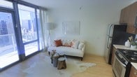 This single-floor space showcases a thoughtfully designed living area that balances modern architecture with warm, inviting décor. The room is anchored by large floor-to-ceiling windows dressed with sheer white curtains, allowing abundant natural light to fill the interior while offering views of the surrounding urban environment. The light hardwood flooring runs throughout, contributing to an open and airy atmosphere.

A tufted cream sofa serves as the focal point of the living area, styled with coral and floral accent pillows that introduce warmth and color against the predominantly white palette. Rustic wooden stump side tables and a neutral cowhide-style area rug add an eclectic, nature-inspired contrast to the clean, contemporary surroundings. A tall arc floor lamp provides additional ambient lighting, while a framed artwork piece adorns the smooth white wall, lending a refined finishing touch.

The overall aesthetic is bright and minimalist, with carefully chosen furnishings that create a welcoming and stylish environment. The seamless flow of natural light, neutral tones, and curated décor elements come together to define a space that feels both functional and visually cohesive.