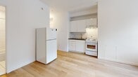 Located at 1240-1250 Rue du Fort in Montréal's vibrant urban core, this studio unit offers a thoughtfully organized single-floor layout designed to maximize every inch of available space. The interior is characterized by a clean, all-white palette with light hardwood flooring that runs throughout the main living area, creating a bright and cohesive atmosphere.

The compact kitchen is efficiently appointed with white cabinetry, a dark-countertop sink with a brass-toned faucet, an electric range with overhead ventilation, and a full-sized refrigerator. Upper and lower cabinet storage ensure practical organization within the streamlined space.

The main living area benefits from a window that invites natural light and frames views of the surrounding neighbourhood. A built-in wall unit featuring upper cabinets, open shelving, and lower drawers provides ample dedicated storage without encroaching on the open floor plan. A baseboard heating unit runs along the exterior wall, ensuring year-round comfort.

The bathroom is finished in crisp white tile, featuring a full soaking tub with an overhead shower, a vanity with under-sink storage, and a large mirror. White floor tiles complete the clean, functional aesthetic.

An entry corridor with a coat rod and thermostat control panel offers a welcoming transition into the unit. Overall, the space presents a practical and well-lit environment suited for efficient urban living in Montréal.