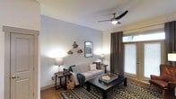 This single-floor space offers a thoughtfully designed interior that balances comfort and modern style. The living area serves as the heart of the home, featuring smooth, light-painted walls that create a clean and airy atmosphere. A sleek ceiling fan with integrated lighting anchors the room from above, enhancing both airflow and ambiance. Natural light filters in through a large window dressed with elegant dark curtains and horizontal blinds, offering both brightness and privacy as needed. The living room is appointed with a plush, neutral-toned sofa accented by decorative throw pillows and a cozy green blanket, paired with a bold dark wood coffee table that grounds the space. A patterned area rug adds visual texture underfoot, complementing the warm hardwood flooring beneath. Side tables with ambient lamps provide soft, layered lighting throughout the room. The walls are tastefully decorated with geometric gold wall shelves and a framed botanical print, adding personality without overwhelming the space. The overall ambiance is warm, inviting, and well-curated, making it an ideal setting for relaxed everyday living.