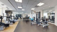 This modern fitness center is thoughtfully designed to support a comprehensive workout experience. The space is organized into distinct zones, seamlessly transitioning between cardio, strength training, and free weight areas. A raised platform with patterned flooring houses a row of cardio machines, including treadmills, ellipticals, and a stationary bike, all positioned to take advantage of the large floor-to-ceiling windows that frame expansive city and outdoor views. Natural light fills the space, creating an energizing atmosphere for exercise.

The strength training area features a multi-station cable machine, a weight bench, and a well-stocked dumbbell rack with a full range of free weights. Stability balls add versatility to the workout options available. Full-length wall mirrors run the length of the space, amplifying the sense of openness and allowing for proper form checks during exercise.

The fitness center is finished with a drop panel ceiling fitted with recessed and panel lighting that maintains consistent, even illumination throughout. Durable rubber flooring in the open training areas ensures safety and comfort. Large glass doors connect the gym to an adjacent outdoor terrace, offering views of a pool area and open sky beyond, making this facility both functional and inspiring.