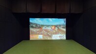 This single-floor space is thoughtfully designed as a premium indoor golf simulator suite that seamlessly blends athletic functionality with upscale lounge aesthetics. The space is divided into two distinct zones: a simulator bay and a comfortable social area.

The simulator bay features a Golfzon Vision+ system, an award-winning golf simulator recognized as a top-rated platform from 2017 to 2020. The bay is outfitted with artificial turf flooring, padded black wall surrounds, and a large projection screen, creating an immersive environment for virtual play. Bean bag chairs are positioned along the turf for spectators, adding a casual viewing option within the bay.

The adjacent lounge area is finished in deep charcoal tones with high ceilings that lend a dramatic, intimate ambiance. A large L-shaped leather sectional sofa anchors the seating area around a live-edge wood coffee table. Decorative pendant lights hang overhead, complementing a bold colorful artwork on the wall. A built-in wet bar with a beverage cooler and wood cabinetry provides convenient refreshment access. Floor-to-ceiling curtains, textured wall panels, and teal accent seating complete the sophisticated, moody interior design. A ceiling-mounted projector and wall-mounted flat-screen TV serve dual entertainment purposes throughout the space.