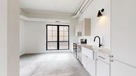 Located at 120 Third Ave N in Minneapolis' vibrant North Loop neighborhood, this one-bedroom, one-bathroom unit showcases a clean industrial-modern aesthetic throughout. Polished concrete floors span the entire space, complementing the exposed ductwork and white-painted structural elements that reference the building's industrial heritage.

The open-concept living and kitchen area is anchored by bold, black-framed grid windows that flood the space with natural light and offer views of the surrounding urban streetscape. The kitchen is well-appointed with white and warm-toned cabinetry, a classic farmhouse sink with a matte black faucet, white subway tile backsplash, and a full suite of stainless steel appliances, including a range, dishwasher, and refrigerator. A wall-mounted industrial-style sconce adds a stylish finishing touch.

The bedroom is a bright, compact retreat featuring a black-framed single window with views toward the neighborhood and an exposed concrete column that reinforces the loft-inspired character of the unit. A sliding barn door with black hardware adds a space-conscious and design-forward element.

The bathroom maintains the same crisp, minimalist palette with white cabinetry, concrete floors, and a narrow window that brings in natural light. Throughout the unit, the cohesive use of black hardware, white walls, and raw materials creates a refined, contemporary urban living environment.