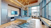 Located at 12004 Inspiration St in Reston, Virginia, this sophisticated amenity lounge is a showcase of contemporary design and premium finishes. The single-floor space is unified by warm light oak hardwood flooring and dramatic double-height ceilings that lend an airy, expansive feel throughout. Floor-to-ceiling glazed walls flood the interior with abundant natural light while framing sweeping views of the surrounding Reston skyline and an adjoining outdoor terrace. A sculptural, large-scale pendant chandelier composed of interlocking geometric rings serves as a striking focal point, anchoring the central lounge and games area. A contemporary pool table with a clean-lined wood frame takes center stage atop a textured area rug, complemented by bar-height seating along the window line. A built-in wet bar and kitchenette, featuring stainless steel appliances and a wall-mounted flat-screen TV set against warm wood cabinetry, adds functional elegance. A separate lounge nook offers a large-format display wall, built-in shelving with curated greenery, and plush seating with direct sightlines to the outdoor terrace. Additional banquette and high-top seating areas provide versatile gathering spaces, while glass-partitioned rooms offer more intimate settings. The overall ambiance is modern, refined, and inviting.