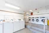 Located at 200 Bay St S in Hamilton, Ontario, this shared laundry facility offers a clean, functional, and well-organized environment designed to accommodate high-volume use. The single-floor space is divided into two interconnected areas, each purpose-built to support an efficient laundry experience.

The main laundry area is bright and welcoming, featuring large windows that allow generous natural light to fill the room and offer views of surrounding greenery. The floor is finished with light-toned ceramic tiles throughout, contributing to a clean and contemporary aesthetic. White gloss wall tiles, accented by a horizontal metallic border stripe, line the walls and add a polished, hygienic finish.

Multiple front-loading washing machines and stacked dryer units are arranged in rows, providing substantial capacity for residents. A long granite-topped folding counter runs along one wall, offering ample surface space for sorting and folding laundry. The space also includes a soap dispenser station and payment terminals conveniently positioned near the machines.

Safety and maintenance are well considered, with a fire extinguisher, sharps waste container, and clearly posted signage throughout. The facility is accessed via a railed entryway with steps, and a utility room is accessible from the main floor. Overall, this laundry facility combines practicality with a clean, modern design to serve its building residents effectively.