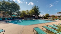 Located at 100 Hunsberger Dr in Limerick, Pennsylvania, this outdoor amenity area serves as a premier recreational destination within the community. At its center sits a generously sized swimming pool with clear, inviting blue water, designed with a curved layout that encourages both lap swimming and leisure. Adjacent to the main pool is a dedicated hot tub spa, finished with tiled stone detailing and seamlessly integrated into the surrounding deck. The expansive concrete pool surround provides ample space for lounging, lined with teal chaise loungers and shaded by several patio umbrellas for comfortable outdoor relaxation. A charming white gazebo anchors one corner of the deck, offering a shaded seating area ideal for gathering or unwinding away from the sun. Mature trees border the fenced perimeter, providing natural privacy and a lush, park-like backdrop. The adjacent clubhouse building features a broad wall of windows and a cupola roofline, allowing natural light to fill the interior while maintaining a visual connection to the outdoor space. A gas grill is also available on the deck, adding convenience for outdoor entertaining. The entire area is enclosed by a white perimeter fence, ensuring a safe and well-defined recreational space open to the sky above.