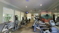 This fitness center is thoughtfully designed to support a full range of workout routines. The space is bathed in natural light, thanks to multiple large windows fitted with roller shades, creating a bright and energizing environment. Crown molding and recessed lighting along the ceiling add a polished, refined touch rarely found in gym settings.

The cardio area is well-stocked with a lineup of treadmills and stair climbers from reputable brands, including True Fitness, providing members with excellent options for aerobic exercise. Complementing these are a variety of strength and resistance training machines, including cable machines and seated resistance equipment, catering to both beginners and seasoned athletes.

A wall-mounted flat-screen television keeps members entertained during workouts, while a striking large-scale mural of an ocean wave adds a dynamic and motivating focal point to the room. An additional display screen is mounted near the entrance, likely for fitness center rules or announcements.

The flooring features a durable, patterned carpet tile suited for a high-traffic fitness environment. A ceiling fan aids in air circulation, contributing to overall comfort during intense workouts. The curved wall layout gives the space an open, flowing feel, making it both functional and visually appealing.