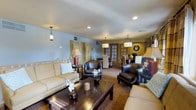 This single-floor suite offers a well-appointed living space characterized by a warm and inviting ambiance. The main living area is generously sized, furnished with a pair of cream-toned sofas and dark leather armchairs arranged around a rich, dark wood coffee table. Recessed lighting and pendant fixtures illuminate the space, complementing the neutral wall tones of soft blue and warm yellow. Decorative accents, including floral arrangements and vases, add a refined touch throughout.

A notable rustic element is the large wood-paneled decorative screen, which adds texture and visual interest to the living area. The carpeted flooring in a warm earth tone contributes to the cozy atmosphere. Towards the rear of the suite, French doors with glass panes lead to a separate dining area, where a dark wood dining table and high-backed chairs provide a more formal setting. Pendant lighting above the dining table reinforces a cohesive design scheme throughout the suite. The overall layout is open and flowing, making the space feel both functional and comfortable for relaxed living and entertaining.