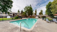 Located at 6506 Doolittle Ave in Riverside, California, this outdoor community amenity area is centered around a generously sized rectangular swimming pool with clear turquoise water, ideal for recreation and relaxation. The pool deck is finished in concrete and provides ample space for lounging, with multiple chaise lounge chairs and side tables arranged along the perimeter for residents to enjoy the Southern California sunshine. A separate circular spa/hot tub is positioned adjacent to the main pool, offering a dedicated space for relaxation. Shaded outdoor dining areas featuring red patio umbrellas and table-and-chair sets provide comfortable gathering spots for socializing. The space is enclosed by a black metal fence for security and safety, while lush mature trees — including large deciduous varieties — surround the area, offering natural shade and a green, park-like atmosphere. A single-story clubhouse or amenity building with a gabled roof, neutral exterior, and red-accented entry doors is situated at the edge of the pool area, likely housing restrooms or community facilities. Well-maintained landscaping with shrubs and planters adds a polished, welcoming character to this community outdoor retreat.