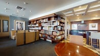 Located at 65 N Madison Ave in the heart of Pasadena, California, this single-floor community space combines refined design with practical functionality. The main lounge area is a standout, anchored by a dramatic floor-to-ceiling built-in bookcase crafted from rich dark wood, housing an extensive collection of books and decorative objects. Coffered ceiling details and recessed lighting add architectural interest overhead, while dark carpeting grounds the space with a polished feel.

Comfortable seating is thoughtfully arranged throughout, including a neutral-toned sectional sofa, vintage-inspired upholstered armchairs in warm gold tones, and a round wood accent table — creating inviting areas for reading, conversation, or casual gatherings. Large windows dressed with sheer curtains and horizontal blinds line one wall, allowing an abundance of natural light and offering views of the tree-lined Pasadena streetscape outside.

The fully equipped kitchen is a highlight, featuring professional-grade stainless steel Wolf appliances including a six-burner gas range with a commercial hood, wall-mounted oven and microwave, and a stainless steel sink with a dishwasher. Deep espresso cabinetry paired with dark granite countertops and a classic checkered tile floor give this space a contemporary yet timeless character, making it well-suited for catering and event use.